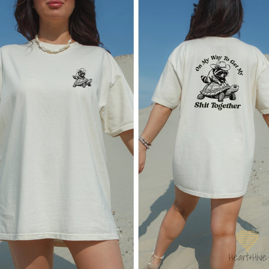 A woman wearing a ivory comfort colors unisex crew neck t-shirt with a graphic of a raccoon and the text 'On My Way To Get My Shit Together' on the front, and the same graphic as a pocket size on the back of the shirt.