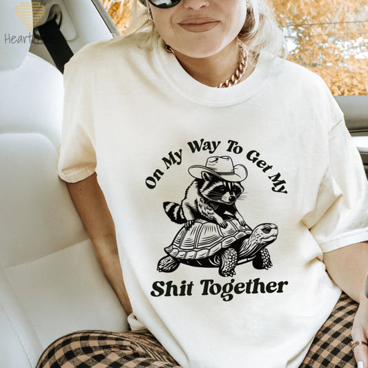 A woman wearing a Ivory Comfort Colors crew neck t-shirt with a graphic print of a raccoon riding a turtle and the text 'On My Way To Get My Shit Together'.