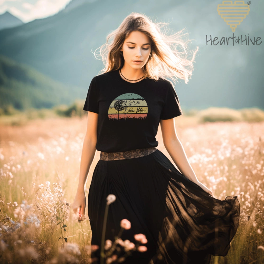 Black tee with a retro sunset circle, grass, and the faded silhouette of a dandelion and 'Blow Me' text.