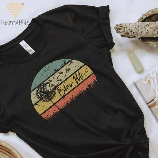 Black tee with a retro sunset circle, grass, and the faded silhouette of a dandelion and 'Blow Me' text.
