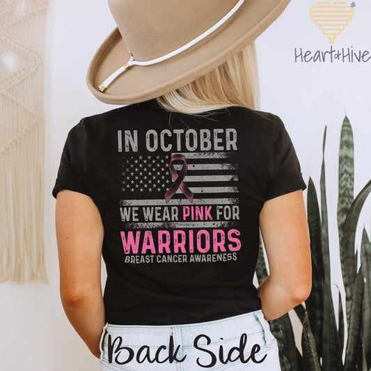 Black tee back features a distressed American flag with a pink ribbon and bold text reading 'In October, We Wear Pink for Warriors – Breast Cancer Awareness' in grey and pink
