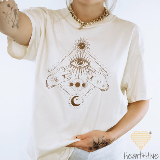 Ivory Comfort Colors tee with a bronze mystical design featuring an all-seeing eye, celestial symbols, and hands adorned with stars and moons.