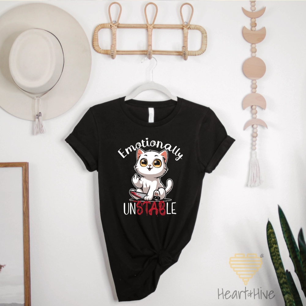 Black tee with a white cat holding a bloody knife with his middle finger up. 'Emotionally' is rounded above and unstable is written below with 'stab' red and dripping.