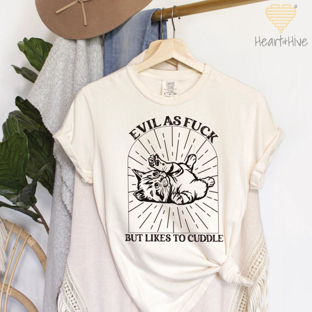 Ivory comfort colors tee with a black line-art illustration of a kitten lying on its back inside a decorative arched frame with radiating lines. The text 'Evil as Fuck but Likes to Cuddle' is placed above and below the design.
