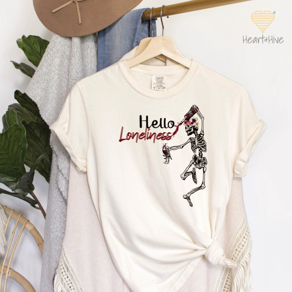 Ivory comfort color tee with a leaping skeleton holding a bottle of wine, splattering red liquid across 'Hello Loneliness' in bold and script fonts.