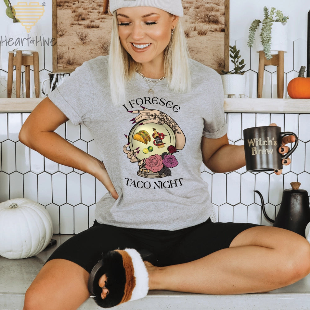 Athletic Heather Grey Tee featuring a mystical crystal ball design. Inside the ball, a taco, lime wedges, and a tequila bottle wrapped by a snake float, framed by tattooed hands with long, dark nails. Surrounded by roses, the text reads, "I Foresee Taco Night".