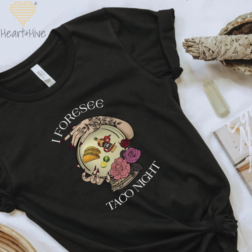 Black tee featuring a mystical crystal ball design. Inside the ball, a taco, lime wedges, and a tequila bottle wrapped by a snake float, framed by tattooed hands with long, dark nails. Surrounded by roses, the text reads, "I Foresee Taco Night".