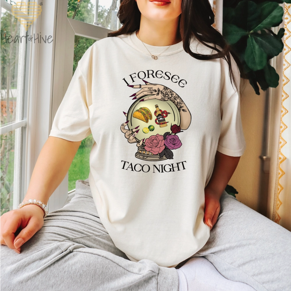 Ivory comfort colors tee featuring a mystical crystal ball design. Inside the ball, a taco, lime wedges, and a tequila bottle wrapped by a snake float, framed by tattooed hands with long, dark nails. Surrounded by roses, the text reads, "I Foresee Taco Night".