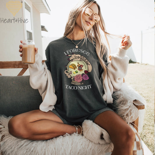 Pepper comfort colors tee featuring a mystical crystal ball design. Inside the ball, a taco, lime wedges, and a tequila bottle wrapped by a snake float, framed by tattooed hands with long, dark nails. Surrounded by roses, the text reads, "I Foresee Taco Night".