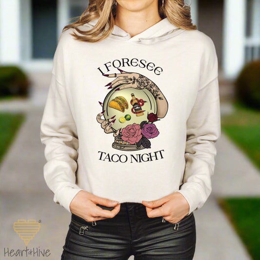 Heather Dust (Beige) cropped hoodie featuring a mystical crystal ball design. Inside the ball, a taco, lime wedges, and a tequila bottle wrapped by a snake float, framed by tattooed hands with long, dark nails. Surrounded by roses, the text reads, "I Foresee Taco Night".