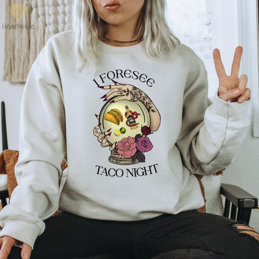 Sand sweatshirt featuring a mystical crystal ball design. Inside the ball, a taco, lime wedges, and a tequila bottle wrapped by a snake float, framed by tattooed hands with long, dark nails. Surrounded by roses, the text reads, "I Foresee Taco Night".