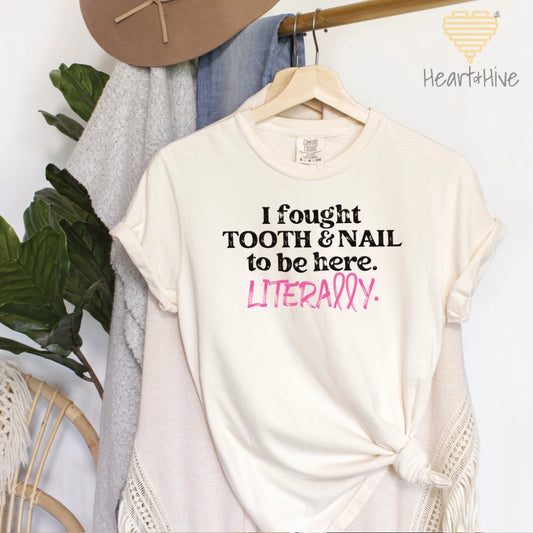 Ivory comfort colors t-shirt with black text reading, "I fought TOOTH & NAIL to be here." The word "LITERALLY" is in pink with a ribbon shape, symbolizing breast cancer survival.