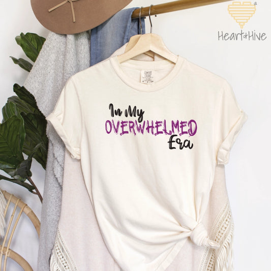 Ivory comfort colors tee featuring the phrase "In My OVERWHELMED Era," with "OVERWHELMED" in purple font and the rest in black.