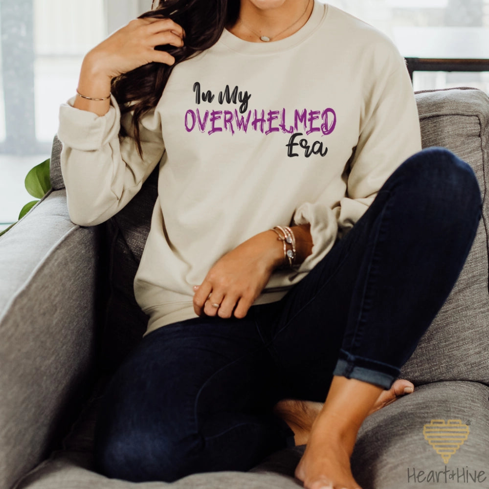 Sand-colored sweatshirt featuring the phrase "In My OVERWHELMED Era," with "OVERWHELMED" in purple font and the rest in black.