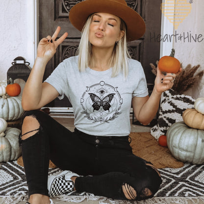 Athletic heather grey tee with a black design of a butterfly, all-seeing eye, crescent moons, celestial symbols, and botanicals.