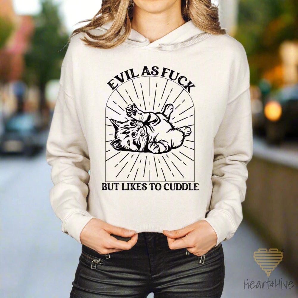 Heather dust (beige) crop hoodie with a black line-art illustration of a kitten lying on its back inside a decorative arched frame with radiating lines. The text 'Evil as Fuck but Likes to Cuddle' is placed above and below the design.
