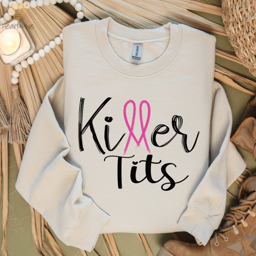 Sand-colored sweatshirt featuring the phrase "Killer Tits" in black script, with the letter "ll" stylized as a pink breast cancer ribbon.
